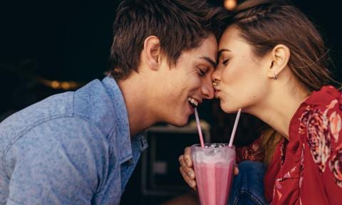 Dos adolescentes dándose un beso mientras beben un batido de fresa.
