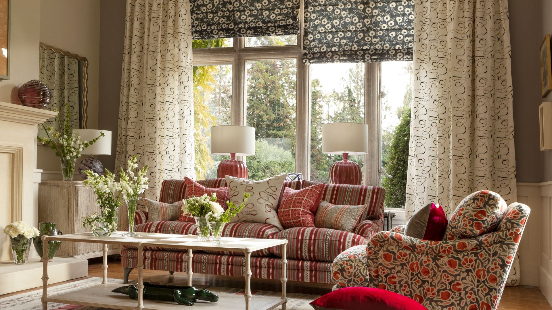 Salón con estores y cortinas de flores, sofá de rayas rojas, sillón de flores, alfombra y cojines en el suelo