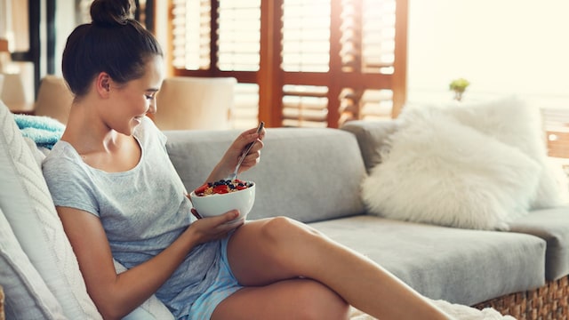 mujer comiendo un bol de yogur y frutos rosos en el sofá de su casa