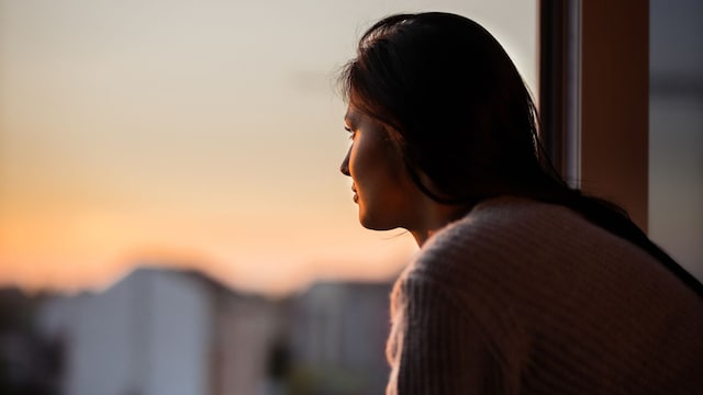 Mujer joven soñando de día mientras mira por la ventana de su apartamento durante la hora del café.