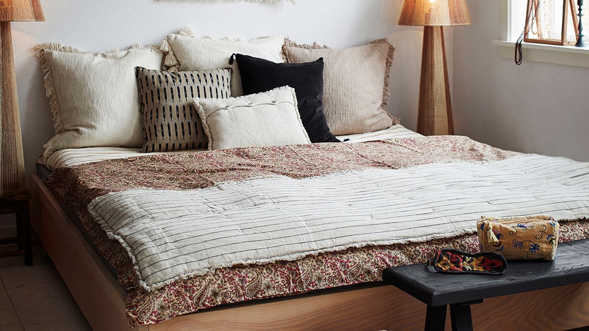 Dormitorio con cama de madera, tapiz en la pared, lámparas de fibras y banqueta a los pies de la cama 