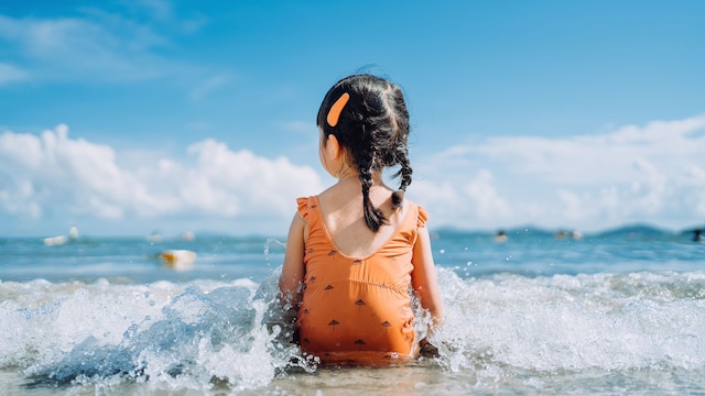 Rear view of lovely little Asian girl sitting by the seashore at the beach and being splashed by waves. Having fun at beach on a sunny Summer day