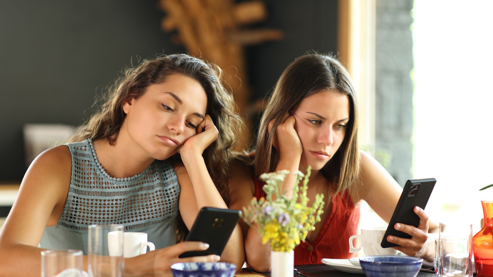 dos chicas jóvenes aburridas en un restaurante mirando sus móviles