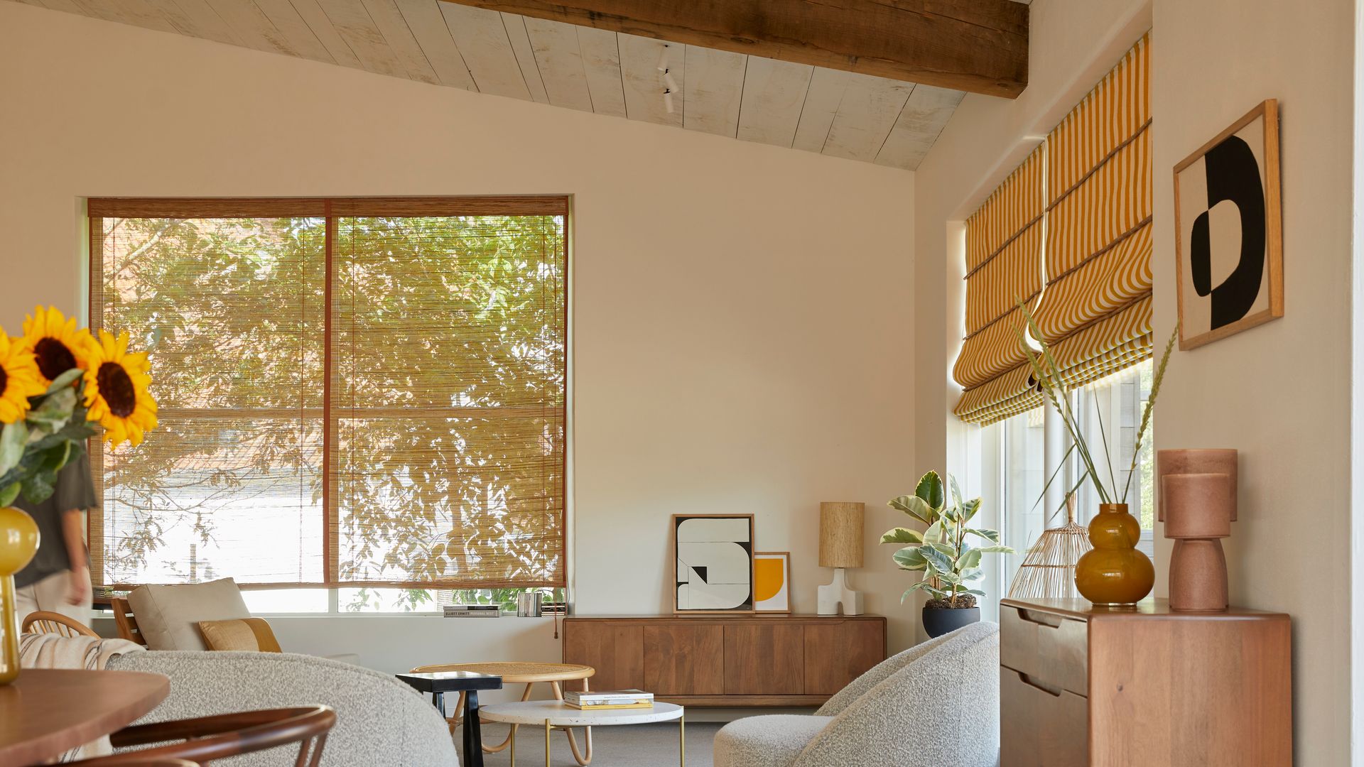 Salón con sofás curvos blanco, aparador y cómoda de madera, jarrón con girasoles, cuadro amarillo y estores de rayas amarillas y blancas