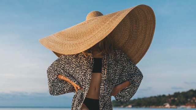 Mujer en la playa con bikini negro, blusa de leopardo y sombrero XXL