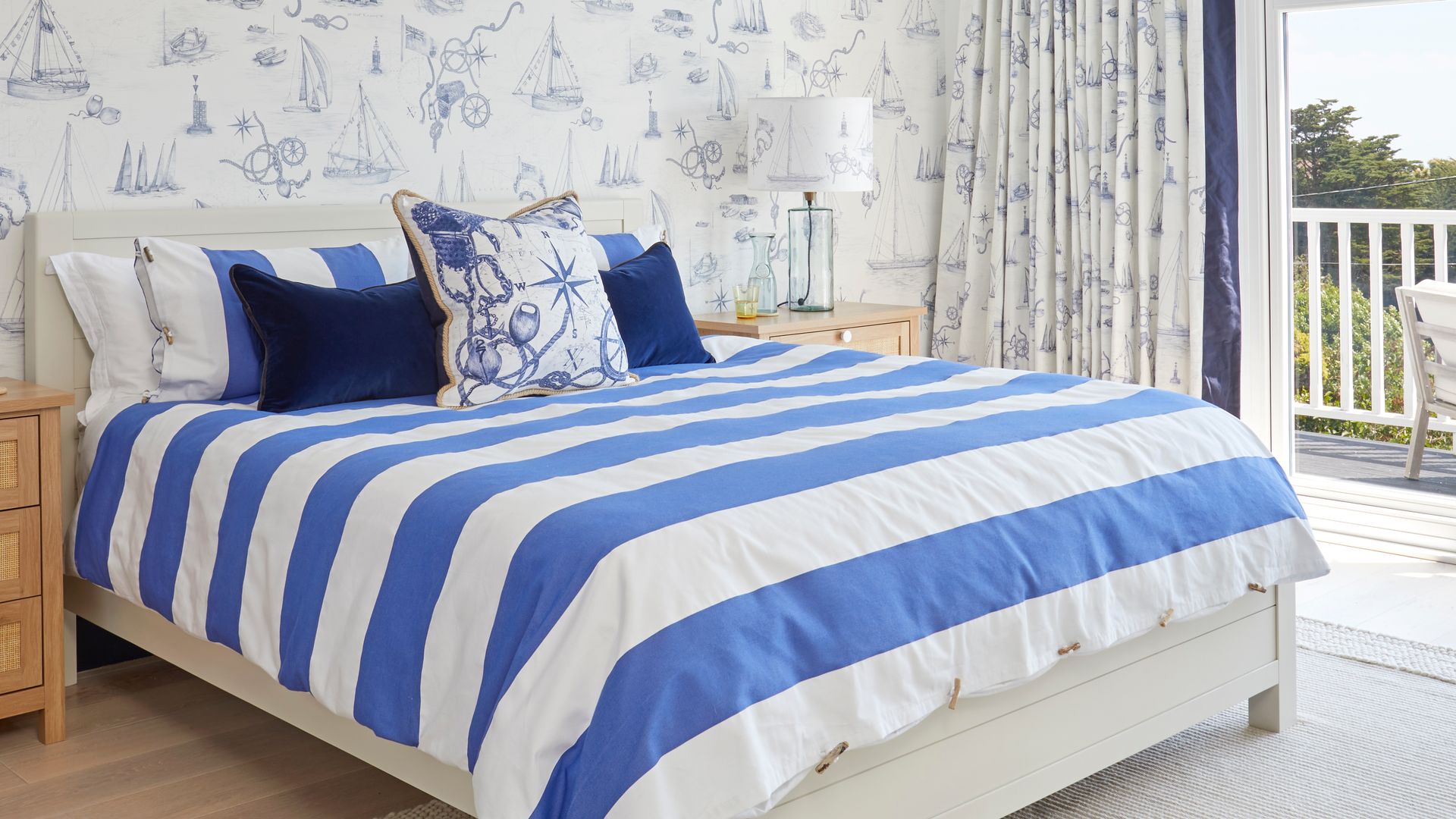 Dormitorio marinero con vigas blancas en el techo, papel pintado azul y blanco con motivos marinos, ropa de cama de rayas azul y blanco, cortinas y salida a la terraza