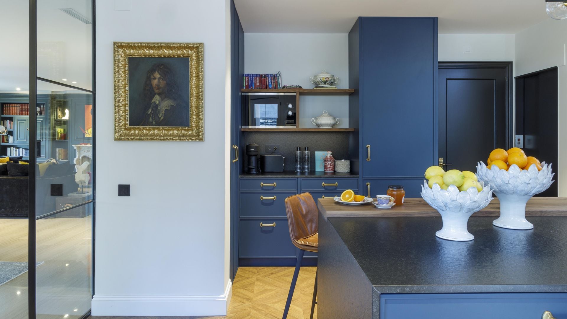 Cocina con muebles azules, cuadro con marco dorado, fruteros blancos y suelo de madera 