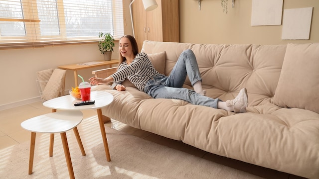 mujer joven tumbada en el sofá comiendo patatas fritas y bebiendo un refresco de cola