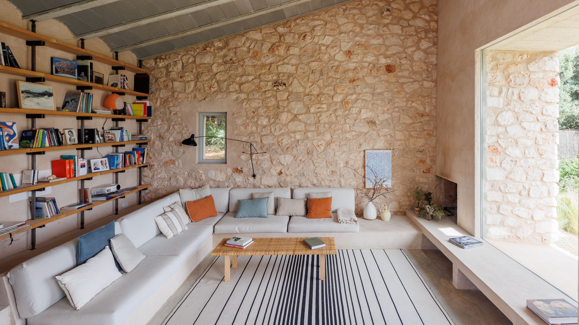 Salón con pared de piedra, estantería de madera, sofá blanco rinconera, gran ventanal y alfombra