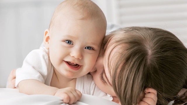 Un bebé sonriendo mirando a cámara mientras su madre le da un beso
