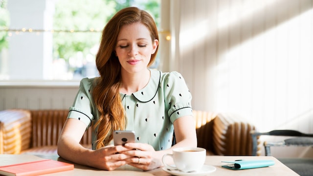 mujer sentada mirando su teléfono móvil con un café en la mesa