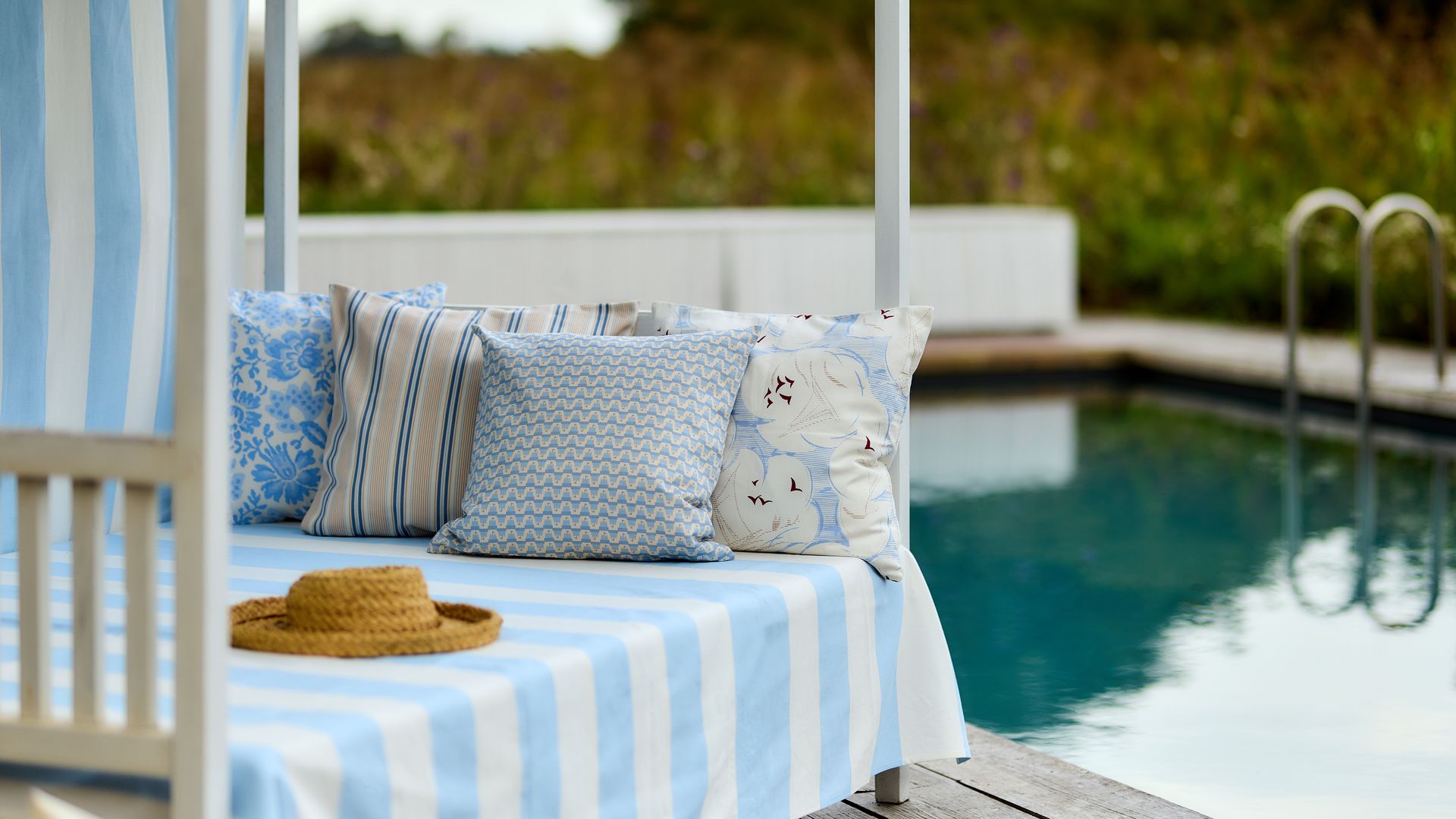 Cama balinesa junto a la piscina con tela de rayas azul y blanca, cojines azules y blancos, sombrero de paja