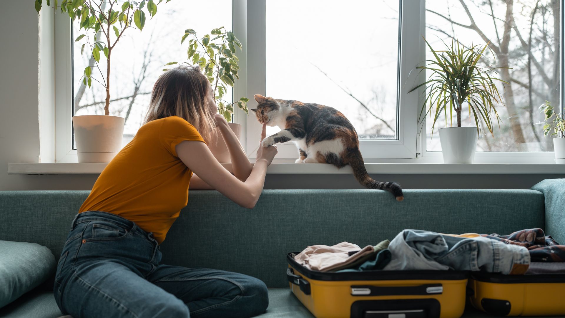 mujer con un gato junto a la ventana, con una maleta abierta sobre el sofá