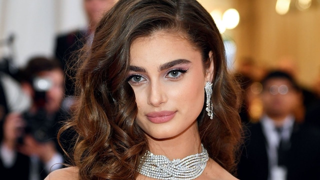 taylor hill posa en la alfombra roja con vestido en rosa palo maquillaje en tonos rosados melena larga ondulada y joyas