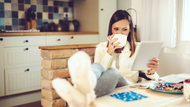 mujer sentada en la cocina de su hogar abrigada con taza de caf en mano y tablet