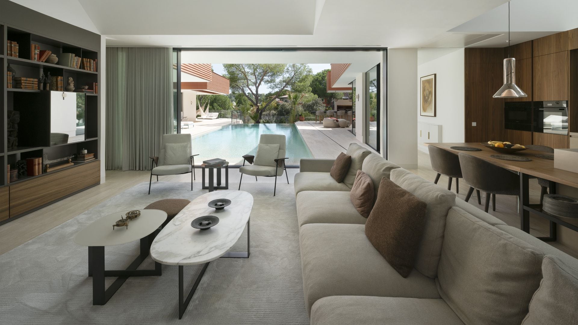 Salón, comedor y cocina abiertos a la piscina mediante un gran ventanal, sofás grises, estantería de madera oscura y mesas de centro 