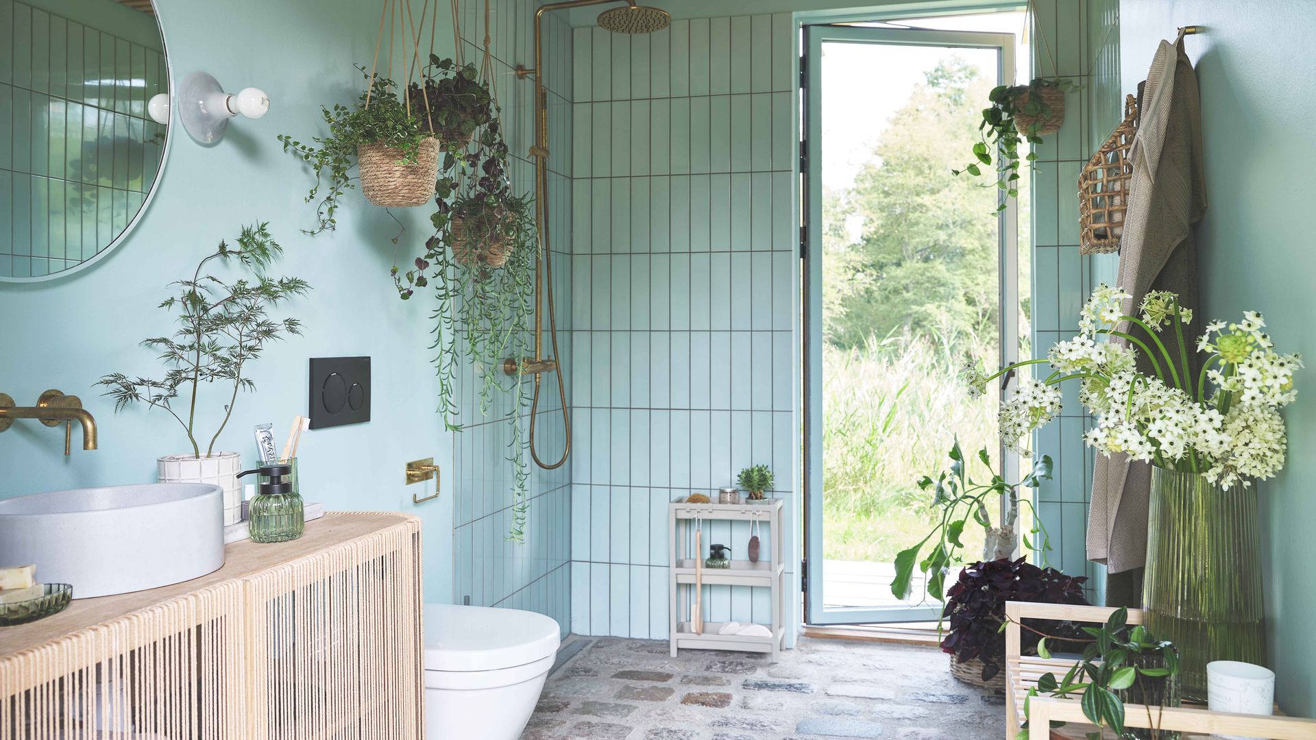 Baño con azulejos azules, suelo de piedra, salida al jardín, mueble de madera, espejo redondo y plantas