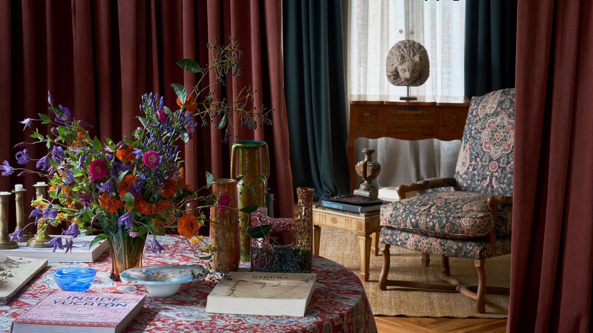 Comedor con cortinas de terciopelo, mesa con mantel estampado, jarrón con flores, libros y adornos