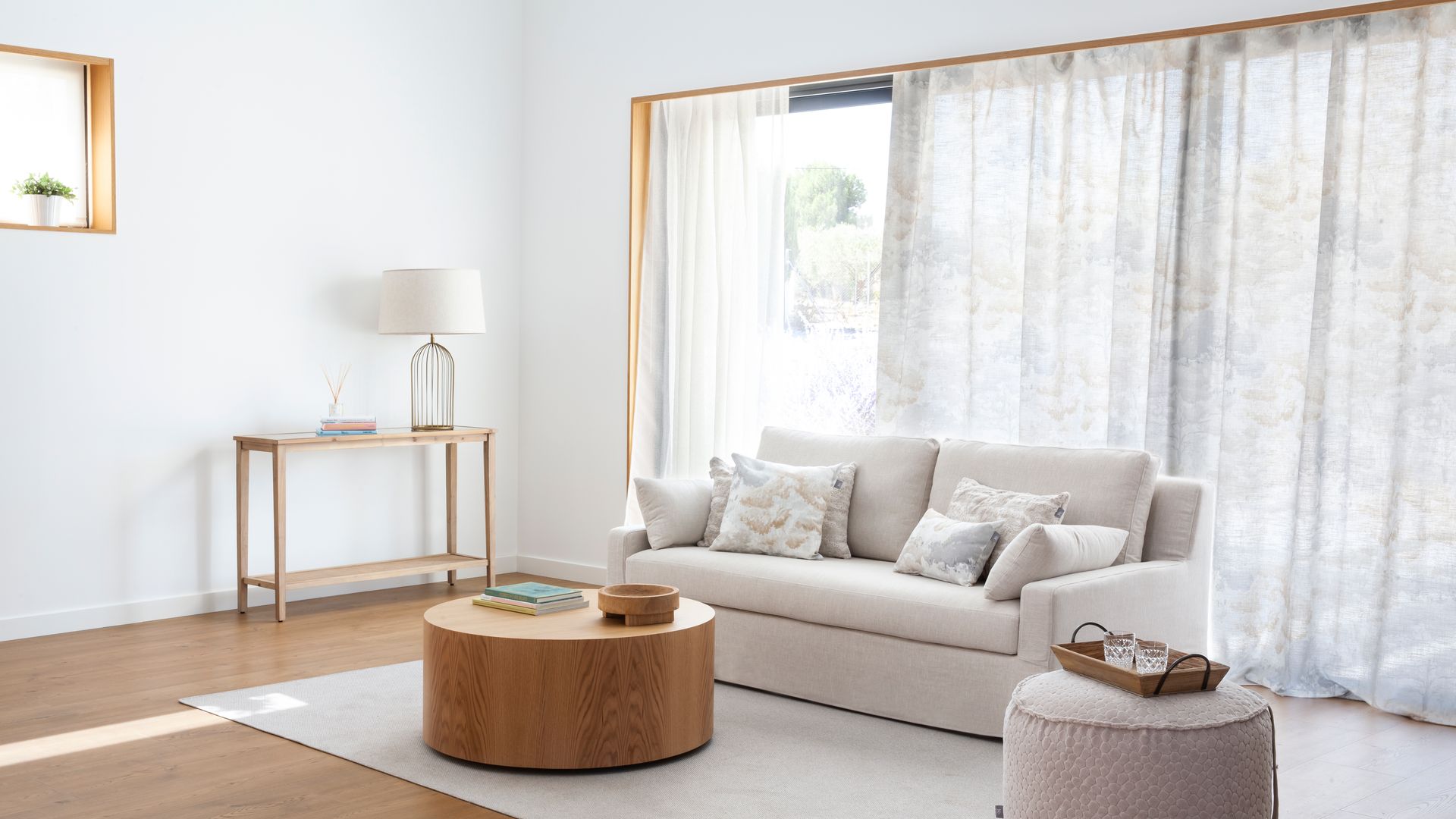 Salón con cortinas y visillos, sofá blanco, mesa de centro de madera y puf