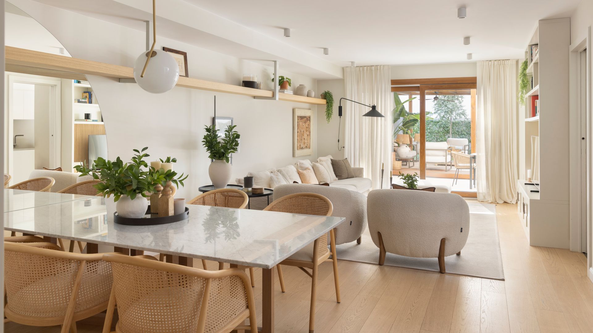 Salón comedor con salida al jardín, suelo de madera, sofá blanco, butacas, alfombra, mesa rectangular, sillas de fibras naturales y lámpara de techo