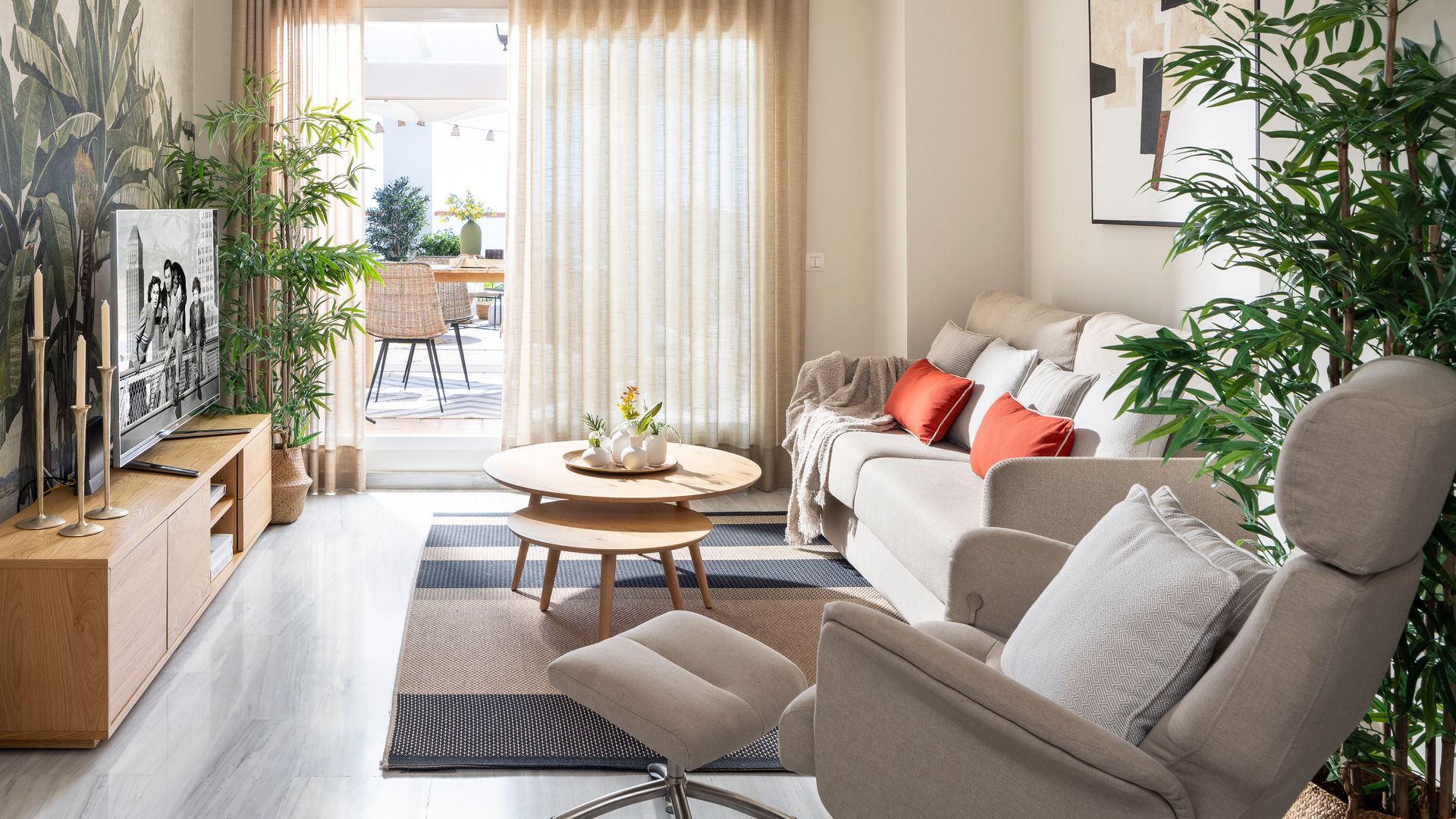 Salón en tonos neutros con papel pintado tropical, mueble de televisión de madera, ventilador de techo y cortinas