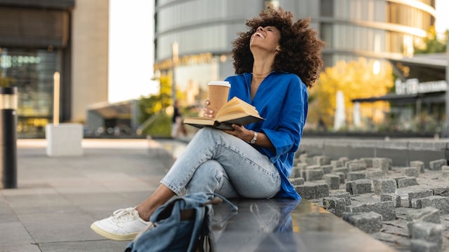 Mujer con un libro y un café en la puerta de la oficina riendo