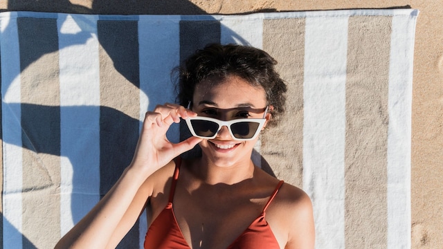Mujer sonriendo con gafas de sol tomando el sol en la playa, encima de la arena