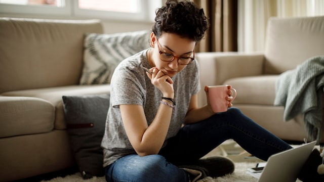 mujer sentada en el suelo de casa, tomando un café mientras trabaja con el portátil