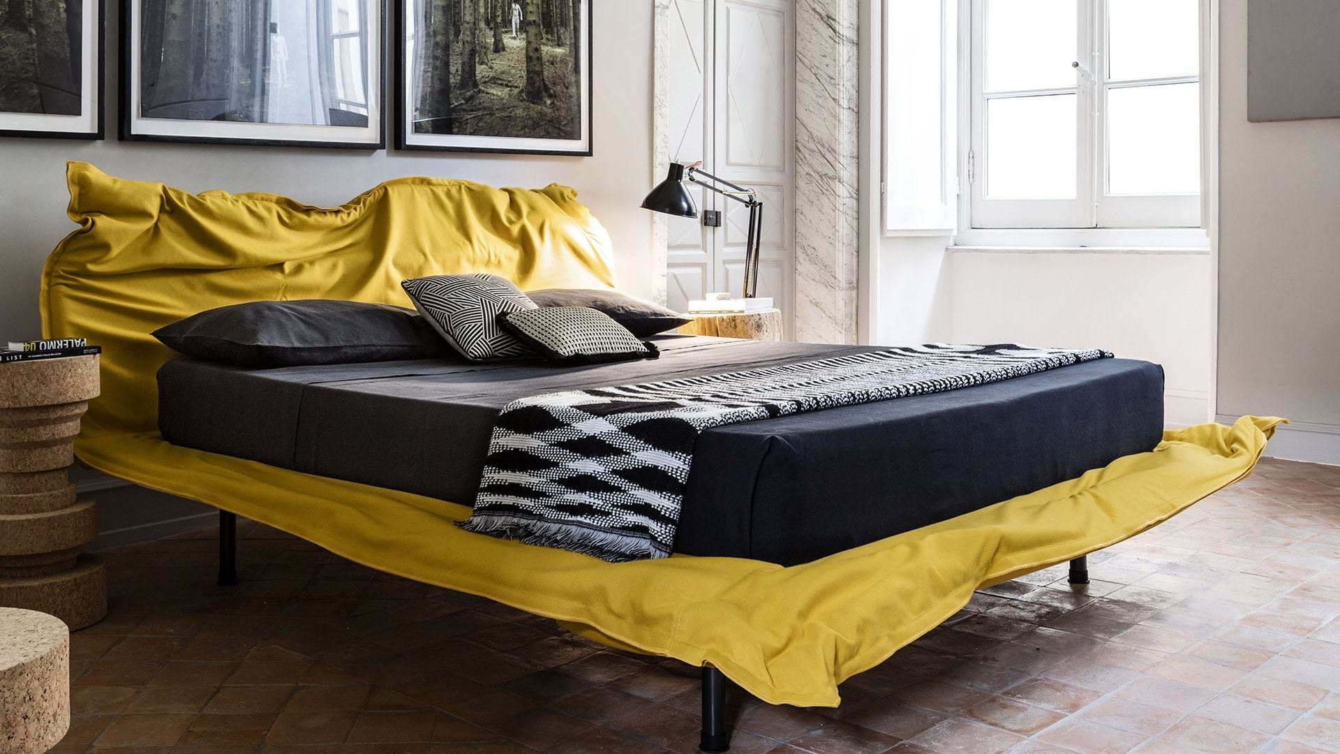 Dormitorio con cama tapizada en amarillo, sábanas negras, fotografías en la pared del cabecero y flexo negro en la mesita de noche 