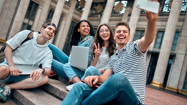 Un grupo de estudiantes está sentado en las escaleras cerca del campus con computadoras portátiles, relajándose, charlando y tomándose selfies con un teléfono inteligente.