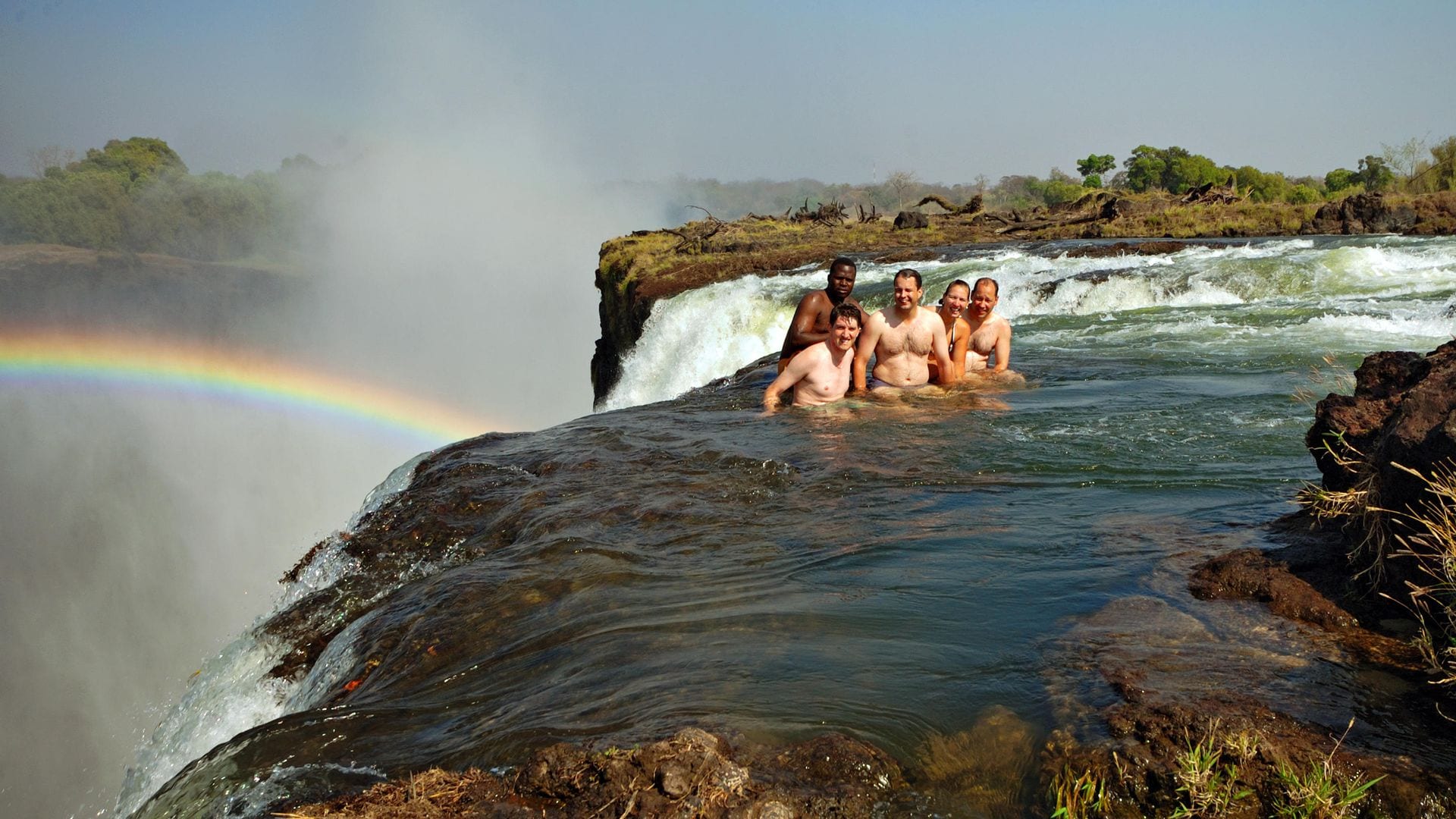 Turistas occidentales con su guía africano nadando justo al borde de las cataratas Victoria, en la llamada Piscina del diablo, en el lado de Zambia