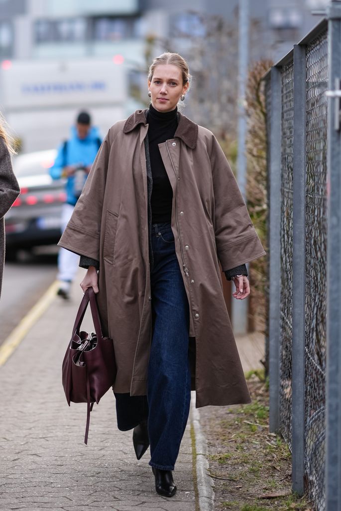 Una invitada lleva pendientes de oro, un cuello alto negro , un abrigo largo marrón , pantalones vaqueros acampanados azules , un bolso de cuero marrón, zapatos de cuero en punta, fuera de Skall Studio, durante la Semana de la Moda de Copenhague AW24 el 31 de enero de 2024 en Copenhague, Dinamarca.