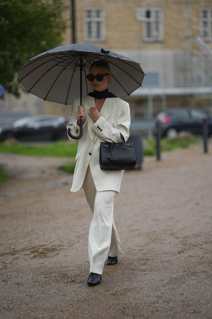 Una invitada lleva un paraguas negro, gafas de sol negras, un pañuelo negro, una chaqueta blazer blanco latte, pantalones de traje blanco latte a juego, un bolso de cuero negro brillante Sac Du Jour de Saint Laurent, zapatos de tacón de cuero negro brillante , fuera de OperaSport, durante la Semana de la Moda de Copenhague Primavera/Verano 2024 el 09 de agosto de 2023 en Copenhague, Dinamarca.