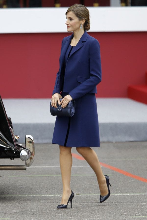 2015: La Reina ha estrenado una chaqueta y vvestido en doble crepe de lana azul marino con cinta en terciopelo azul noche, de Felipe Varela. Llevaba un clutch en piel metalizada azul Prusia con ribetes en cristal de SW, también de Varela. Doña Letizia, que ha recogido su pelo en un sobrio moño bajo, lucía unos pendientes de aguamarinas de Bulgari que la firma le regaló con motivo del nacimiento de la Princesa Leonor y que estrenó en 2006.
