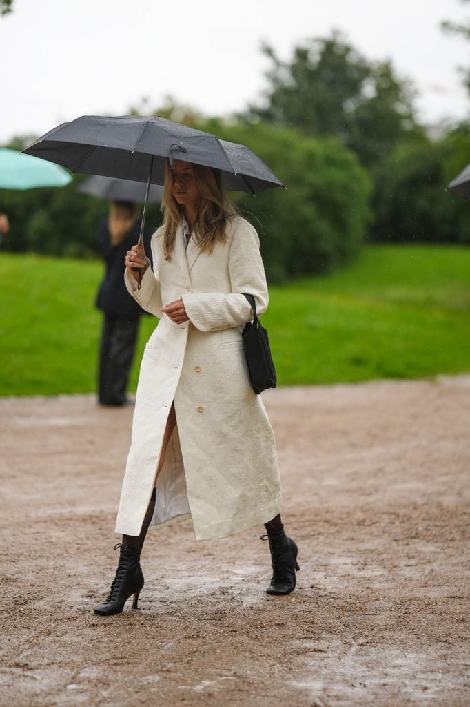 Una invitada lleva un paraguas negro, un abrigo largo blanco café con leche, un bolso de nylon negro, botas / botas altas de tacón de punta con cordones de cuero negro brillante , fuera de OperaSport, durante la Semana de la Moda de Copenhague Primavera/Verano 2024 el 09 de agosto de 2023 en Copenhague, Dinamarca. 