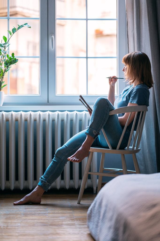 una mujer apuntando datos en su agenda, sentada en una silla y mirando por la ventana