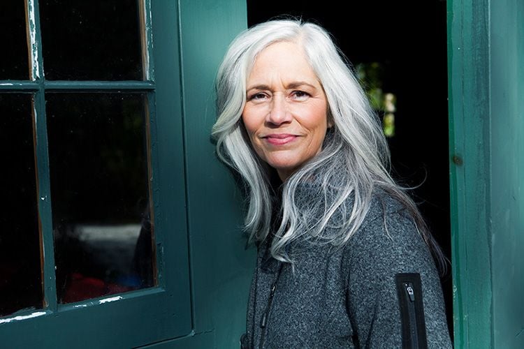 Mujer con el pelo largo de color gris, sontiendo a la puerta de su casa