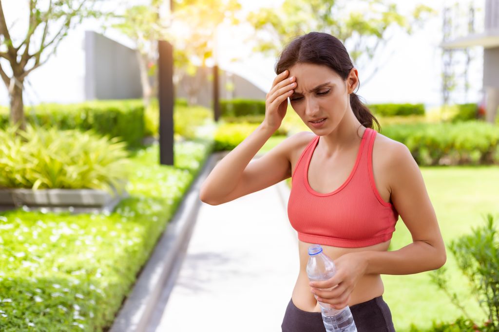 mujer vestida con ropa deportiva, mareada con dolor de cabeza y una botella de agua en la mano