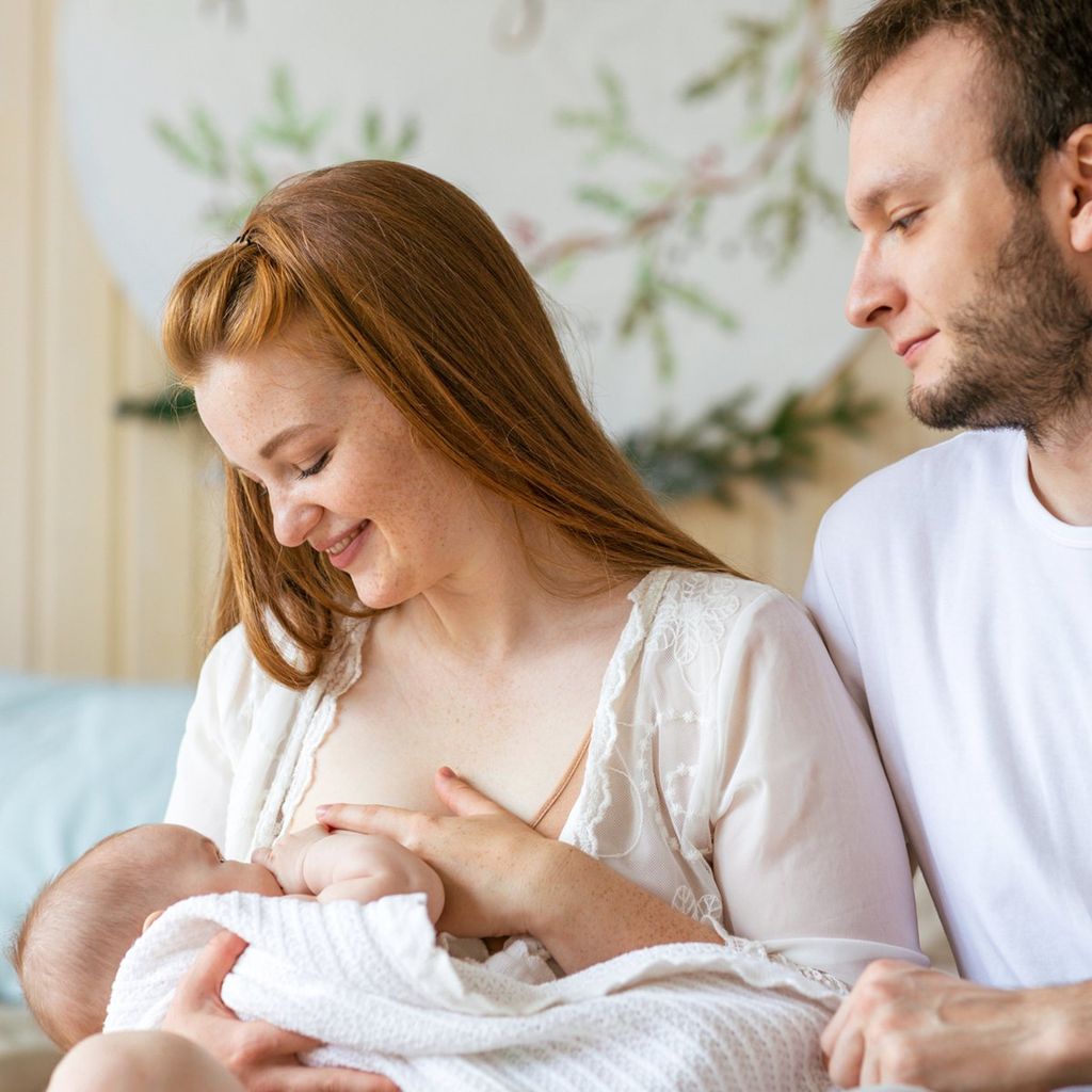 young mother breast feeding her newborn child baby father sits nearby and supports her