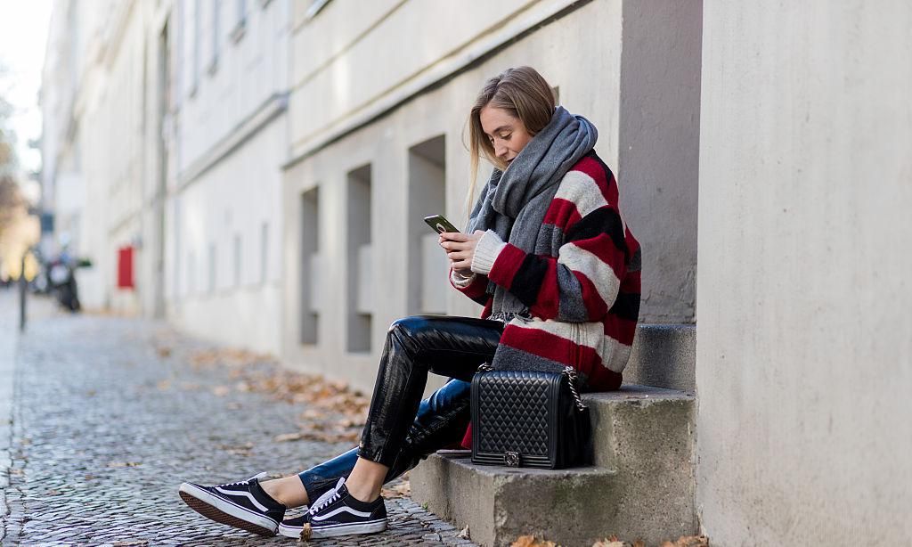 Sonia Lyson sitting on stairs writting a text message wearing a red, white, black grey striped wool coat from Isabel Marant via The Corner Berlin, a grey Acne scarf, black PVC Zara pants, black Vans sneakers via Urban Outfitters, black Chanel bag