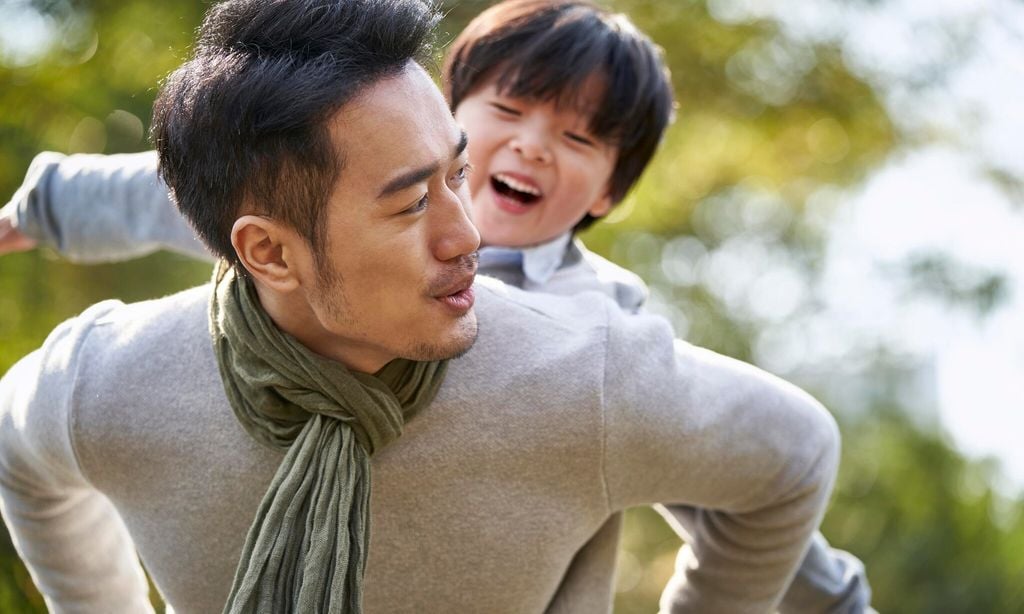 young asian father having fun carrying son on back outdoors in park