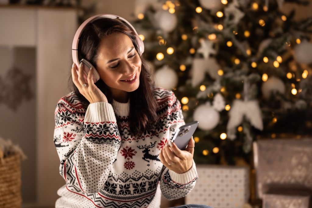 mujer escuchando música con un jersey navideño, junto al árbol de Navidad