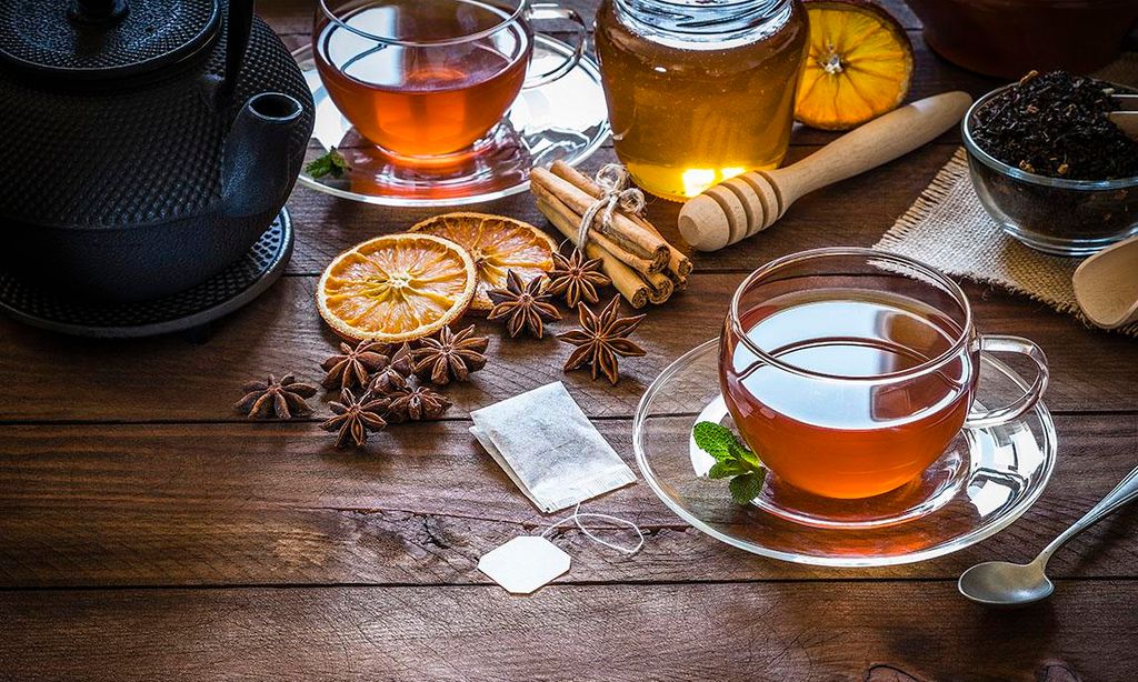 Taza de té llena junto a una cucharadita, una tetera, una bolsita de té y algunos ingredientes como hojas secas de té negro, anís, naranja seca, palitos de canela y un tarro de miel