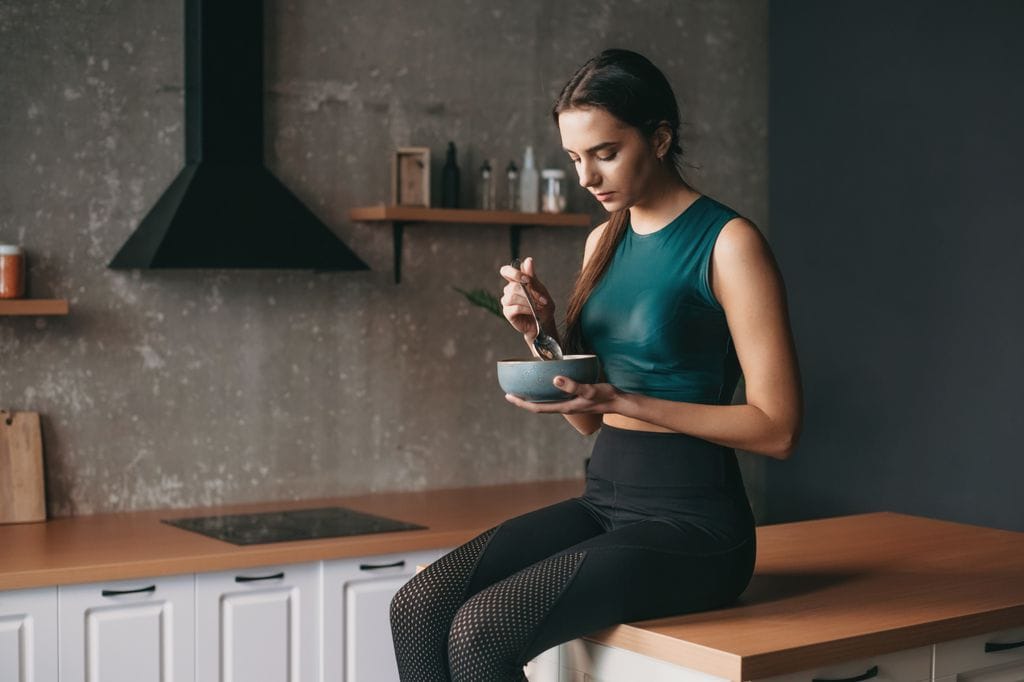 mujer joven con ropa deportiva comiendo sentada en la encimera de la cocina