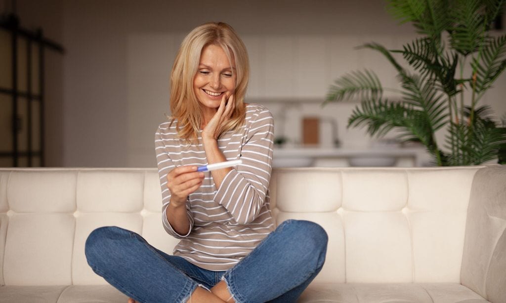 mujer sentada en el sof de su casa feliz con test de embarazo en su mano