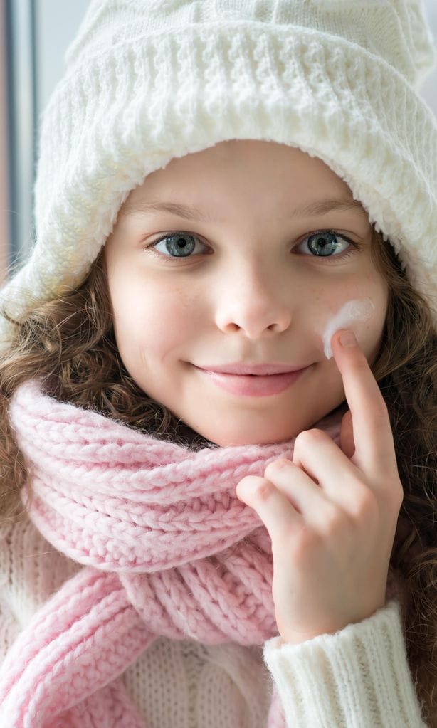 Niña con gorro y bufanda aplicándose crema solar en la cara