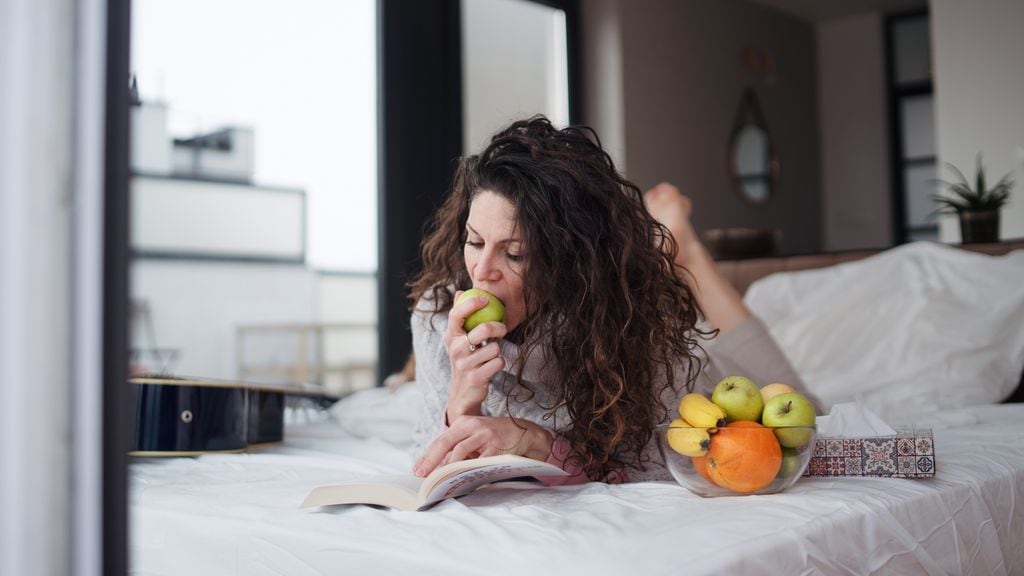 mujer leyendo un libro y comiendo fruta en la cama