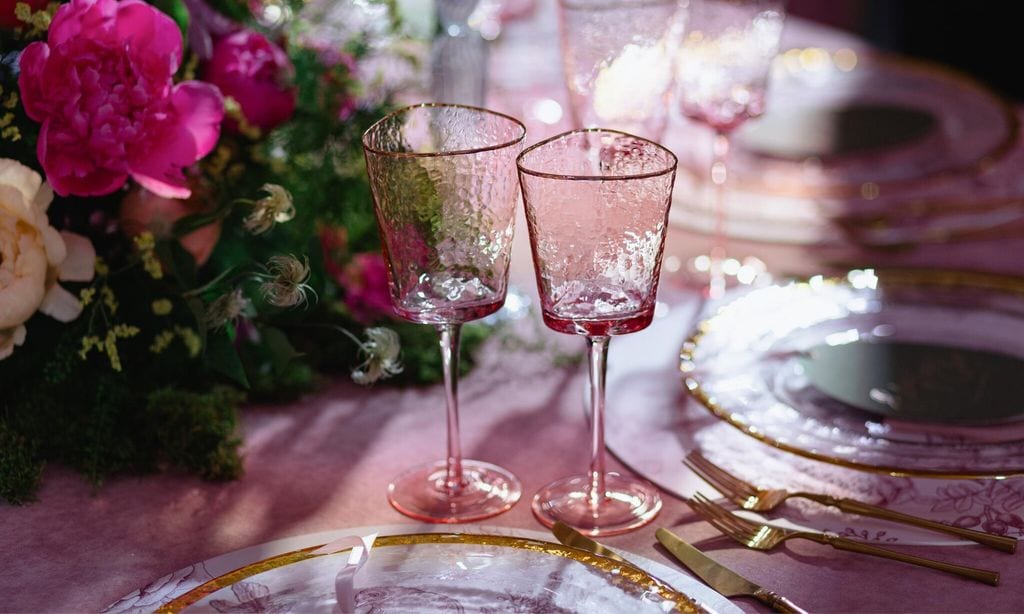 luxurious pink glasses with textured surface are on the festive table it is decorated with fresh bright flowers and greenery golden cutlery are beside the shiny glass plates with golden edge 
