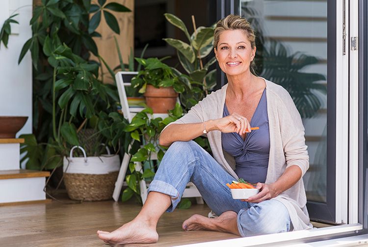 Retrato de una mujer madura sonriente sentada en el suelo con la puerta abierta de la terraza comiendo comida para picar