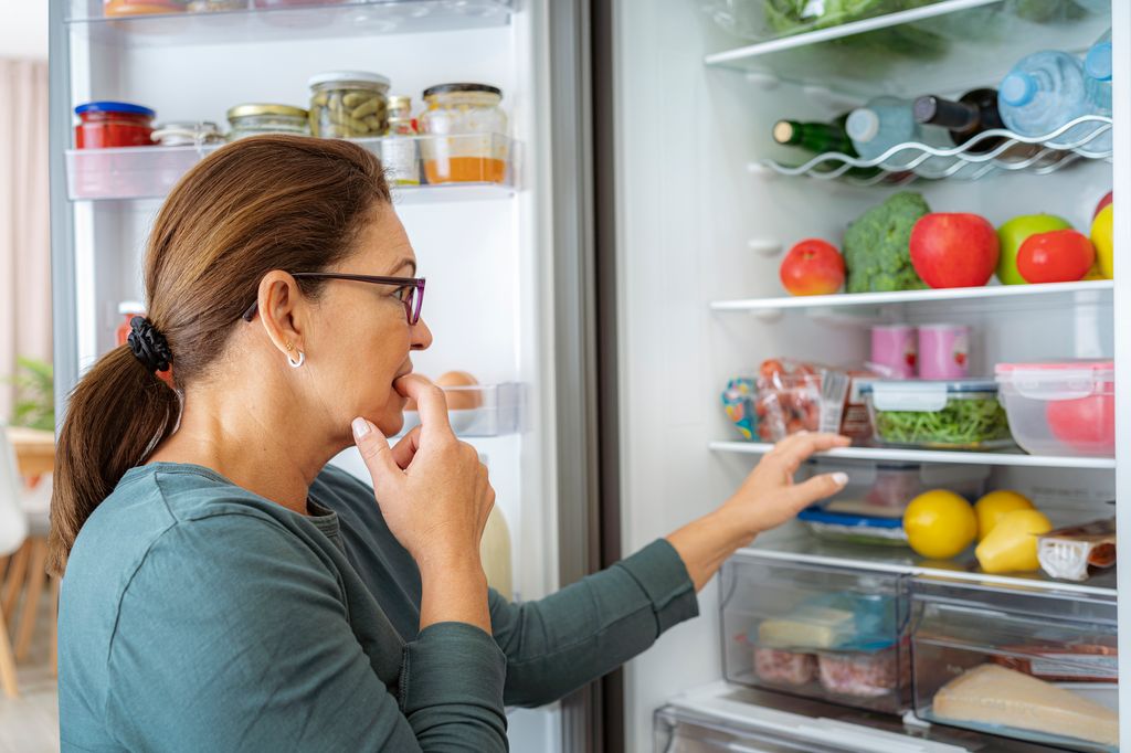 Una mujer dubitativa frente al frigorífico lleno de alimentos